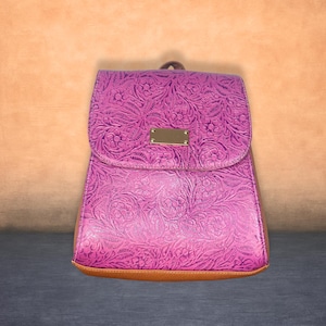 Puede incluir: Una mochila de color morado brillante con un intrincado patrón floral en relieve en toda la superficie. El bolso incluye una placa metálica rectangular de tono dorado en la solapa frontal y una base marrón lisa en la parte inferior.