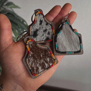 May include: Three cowhide keychains with beaded trim. The keychains are shaped like a cow tag and feature different colored beads. The keychains are brown, white, and gray.