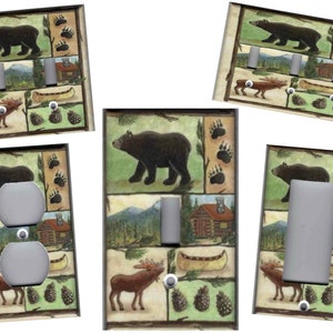 May include: A set of four light switch and outlet covers featuring a woodland theme. The covers feature a black bear, a moose, a cabin, a canoe, pine cones, and paw prints on a green, brown, and beige background.