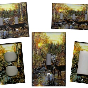 May include: A set of five light switch and outlet covers featuring a scenic image of a forest with two deer standing in a stream at sunset. The image is printed on a white background.