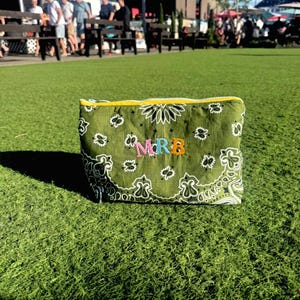 May include: A rectangular olive green pouch featuring a white paisley bandana pattern and a yellow zipper. The letters MRB are embroidered in the center in pink, light blue, and orange thread, creating a personalized look on the fabric surface.