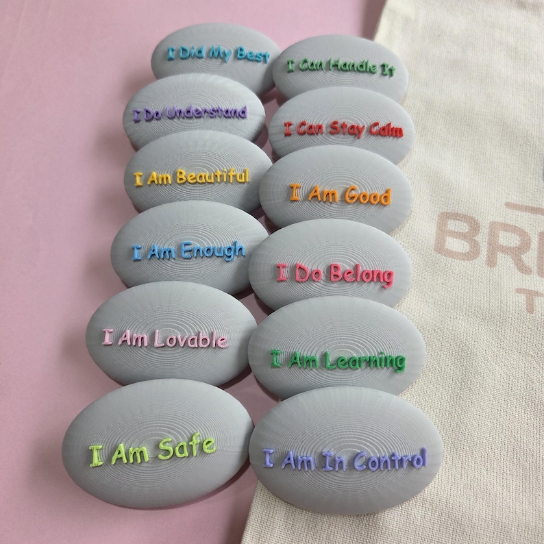 以下が含まれることがあります： カラフルな文字が書かれた灰色の楕円形の石のコレクションです。「私は美しい」や「私は安全です」などの肯定的な言葉が各石に書かれています。石はピンク色の表面に配置されています。