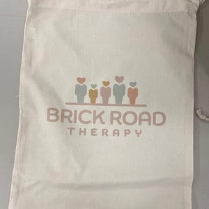 以下が含まれることがあります： クリーム色の巾着袋で、柔らかいピンク色のフォントで「BRICK ROAD THERAPY」の文字が書かれています。テキストの上には、パステルカラーのハート型の頭を持つ5つの図のグラフィックがあります。バッグはテクスチャード加工された生地で作られています。