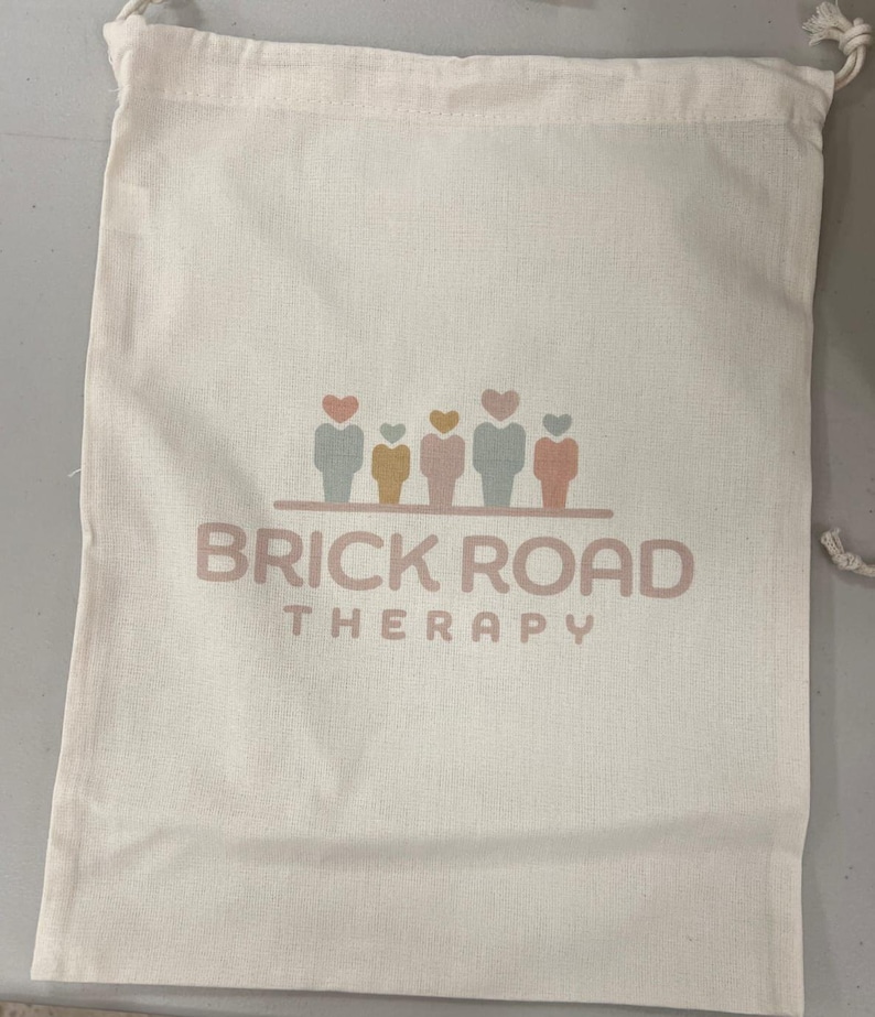 以下が含まれることがあります： クリーム色の巾着袋で、柔らかいピンク色のフォントで「BRICK ROAD THERAPY」の文字が書かれています。テキストの上には、パステルカラーのハート型の頭を持つ5つの図のグラフィックがあります。バッグはテクスチャード加工された生地で作られています。
