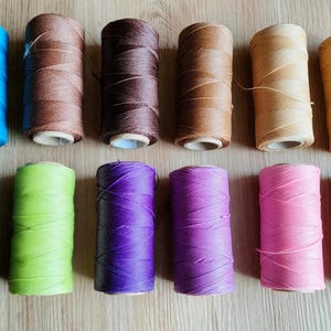 Könnte beinhalten: Eine Auswahl an gewachsten Kordelspulen in verschiedenen Farben: Blau, Braun, Schwarz, Grün, Lila und Rosa. Die Kordeln sind ordentlich auf Pappspulen gewickelt, geeignet für Bastelarbeiten und Schmuckherstellung.
