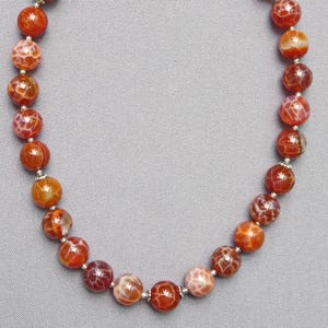 Puede incluir: Un collar con cuentas grandes y redondas en tonos naranja, rojo y blanco. Las cuentas tienen una superficie estampada y están ensartadas con pequeños espaciadores plateados. El collar se muestra sobre un fondo gris.