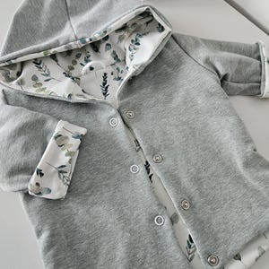 Könnte beinhalten: Graue Kapuzenjacke mit botanischem Print-Futter. Die Jacke hat eine Knopfleiste, lange Ärmel mit umgeschlagenen Manschetten und eine Kapuze. Das Innenfutter und die Manschetten zeigen einen weißen Stoff mit grünen und blauen Blättermustern.