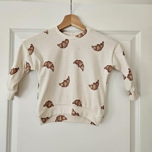 Könnte beinhalten: Cremefarbenes Langarm-Sweatshirt mit Rippstruktur, verziert mit braunen Croissant-Illustrationen. Das Sweatshirt hängt an einem Holzbügel vor weißem Hintergrund. Das Design zeigt verstreute Croissant-Grafiken.