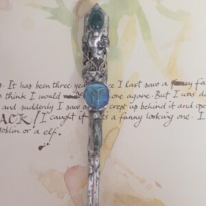 Op de afbeelding: Versierde zilveren pen met een gefacetteerde groene edelsteen en een blauwe gezichtsvormige edelsteen. De pen heeft ingewikkelde details en een goudkleurige punt. De achtergrond bevat handgeschreven tekst en aquarelverf.
