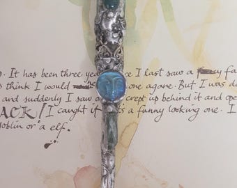 May include: Ornate silver pen with a faceted green gemstone at the top and a blue face-shaped gem. The pen has intricate detailing and a gold-colored tip. The background includes handwritten text and watercolor paint.