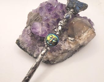 May include: A silver-toned pencil with intricate detailing, topped with a blue butterfly and a round, iridescent face. The pencil rests on a rough, purple amethyst crystal, with a brown crystal in the background. The pencil's tip is a light brown color.