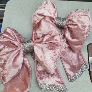 May include: Three large, dusty rose velvet bows with silver glitter accents. The bows have a soft, textured appearance and are arranged on a light green surface. The text "Tel: 0222-499011 ORDER DEPT. Prep 1234" is visible.