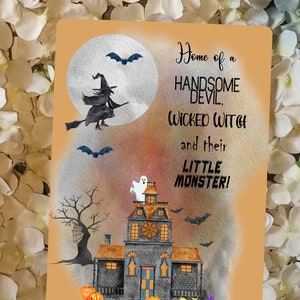 May include: A watercolour illustration of a spooky house with a ghost on the roof, a witch flying on a broomstick in front of a full moon, and the text "Home of a Handsome Devil, Wicked Witch and their Little Monster!"