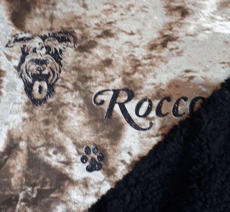 May include: A brown and white patterned dog bed with a black and white dog silhouette, a paw print, and the name "Rocco" embroidered in black.