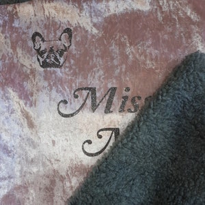 May include: A soft, plush blanket with a brown and white marbled pattern. The blanket has a black faux fur trim and a glittery inscription that reads "Miss A" and a small image of a French Bulldog.