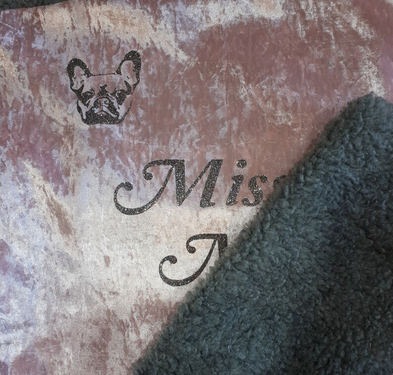 May include: A soft, plush blanket with a brown and white marbled pattern. The blanket has a black faux fur trim and a glittery inscription that reads "Miss A" and a small image of a French Bulldog.