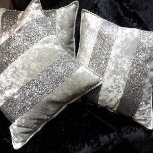 May include: Three silver velvet cushion covers with silver glitter stripes. The cushions are arranged on a black velvet surface.