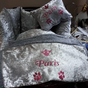May include: A gray velvet dog bed with a gray faux fur trim. The bed has a pink glitter crown and the word "Paris" embroidered on it. There are also pink glitter paw prints on the bed.