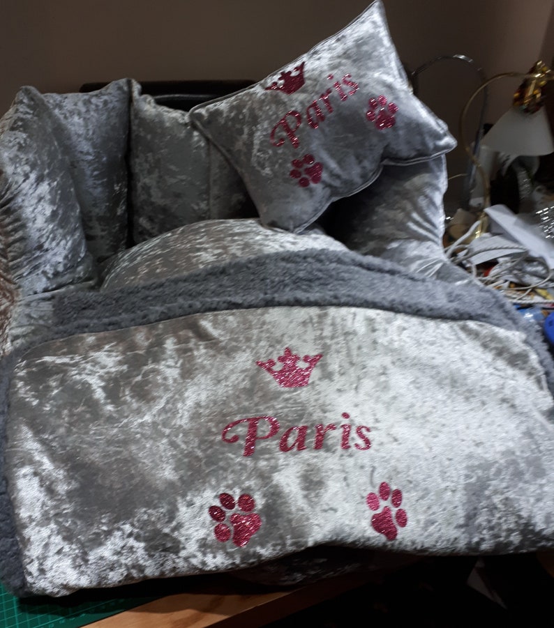 May include: A gray velvet dog bed with a gray faux fur trim. The bed has a pink glitter crown and the word "Paris" embroidered on it. There are also pink glitter paw prints on the bed.