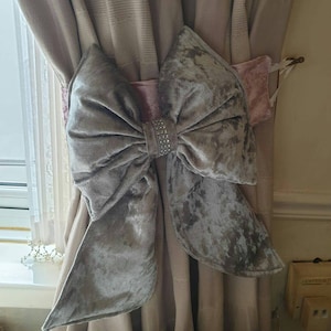 May include: A gray velvet bow with a rhinestone center, used as a tieback for curtains. The bow is attached to a pink velvet ribbon.