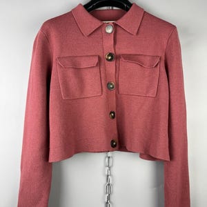 May include: A dusty rose-colored cropped jacket with a collared neckline and two front pockets. The jacket has long sleeves and is fastened with four brown buttons. The garment is hanging on a black hanger.