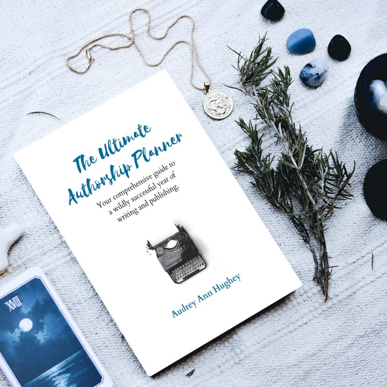 May include: A white book with the title "The Ultimate Authorship Planner" in blue text. The book is lying on a white surface with a sprig of rosemary, a necklace, and a few stones. The book cover also features a vintage typewriter.