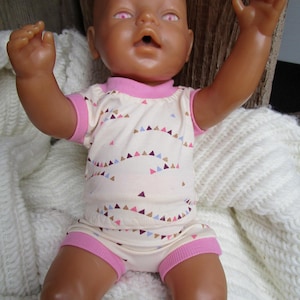 Könnte beinhalten: Eine Babypuppe mit gebräunter Haut und hellen Augen, bekleidet mit einem cremefarbenen Kurzarm-Strampler mit einem Muster aus kleinen, bunten Dreiecken und rosa gerippten Abschlüssen an Hals, Ärmeln und Beinausschnitten, mit dem Datum 26/08/2026 09:00.