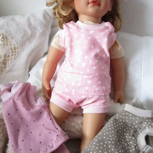 Könnte beinhalten: Eine Puppe mit welligem blondem Haar trägt ein rosa kurzärmeliges Oberteil und eine passende kurze Hose, beide mit weißem Herzmuster. Zwei weitere Puppenkleidungssets sind zu sehen, eines in Mauve mit schwarzen Punkten und eines in Taupe mit weißen Punkten.