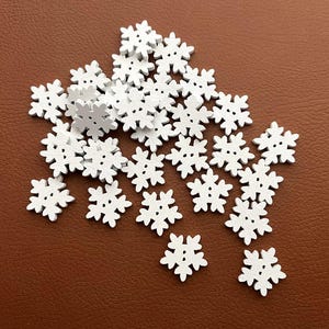 Puede incluir: Un conjunto de pequeños botones blancos de madera con forma de copo de nieve. Cada botón tiene seis puntas y dos agujeros para coser. Los botones están dispersos sobre una superficie marrón, ideales para proyectos de manualidades.