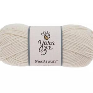 Op de afbeelding: Een bol off-white garen omwikkeld met een papieren label. Het label bevat een bij-icoon met een garenbol als lichaam en de tekst Yarn Bee Pearlspun. Het garen is in een cilindrische vorm gewikkeld met zichtbare vezels over de hele bol.
