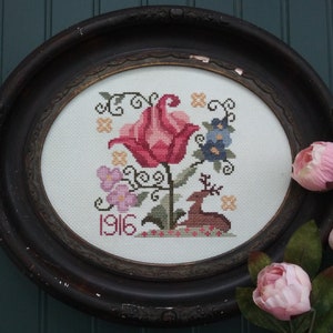 May include: A framed cross-stitch embroidery featuring a pink flower with a deer and the year 1916.