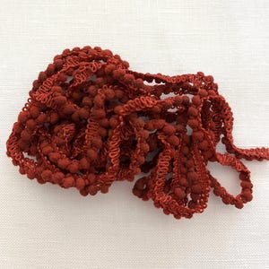May include: A spool of red pom-pom trim. The trim is made of a soft, fuzzy material and has a scalloped edge.
