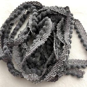 May include: A coiled length of dark gray pom-pom trim featuring a woven fabric header with a repeating zigzag pattern along the edge. The trim is adorned with small, soft, spherical pom-poms attached at regular intervals along the entire length.