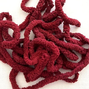 May include: A coiled length of deep burgundy yarn featuring a highly textured, fuzzy, and nubby surface. The yarn is arranged in a loose, tangled pile, showcasing its consistent dark red color and soft, looped construction throughout the strand.