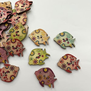 Può includere: Bottoni in legno a forma di pesce con motivi floreali multicolori. Ogni bottone ha fori per cucire. I bottoni sono sparsi su una superficie bianca.