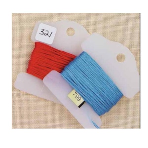 May include: Close-up of embroidery floss in red and blue, with white plastic bobbins. The red floss is labeled "321" and the blue floss is labeled "799". The bobbins have a hole at the top for hanging. The background is a light tan fabric.