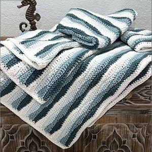 Op de afbeelding: Een handgehaakte deken in wit, lichtblauw en blauwgroen. De deken heeft diagonale strepen en is op een houten oppervlak gevouwen. De randen hebben een witte rand. Een decoratief zeepaardbeeldje staat op de achtergrond.