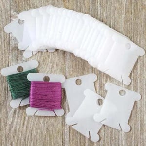 Pode incluir: Uma coleção de bobinas de plástico branco para linha de bordar, algumas vazias e outras enroladas com linha verde e roxa. As bobinas apresentam um formato retangular com entalhes e um orifício circular no topo para organização.