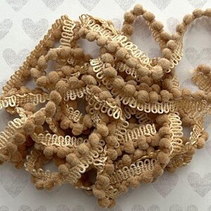 Puede incluir: Una longitud enrollada de cinta de pompones beige con un borde decorativo tejido. El adorno consiste en pequeñas esferas suaves y difusas unidas a un borde de cinta estructurado y con bucles en un color tostado claro a juego, en un montón.