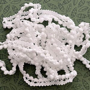 May include: A coiled length of white pom-pom trim featuring a series of small, soft, spherical balls attached to a woven, zigzag-patterned fabric tape. The trim is arranged in a loose, tangled pile, showcasing the uniform white texture.