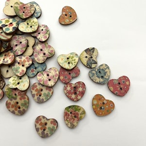 May include: A collection of heart-shaped wooden buttons with floral patterns in various colors. The buttons have two or four holes for sewing and are scattered on a white surface. The floral designs include pink, blue, orange, and green.