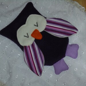 Könnte beinhalten: Lila und weißer Plüsch-Eulen-Spielzeug mit gestreiften Flügeln und einem weißen Gesicht. Das Spielzeug liegt auf einer weißen gehäkelten Oberfläche.