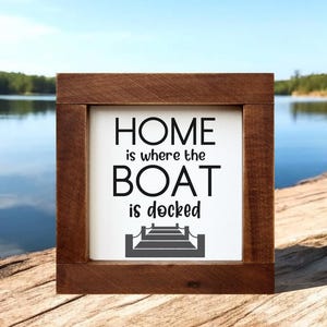 Op de afbeelding: Een vierkant houten bord met een donkerbruine lijst en een wit middenpaneel. De tekst "HOME is where the BOAT is docked" staat in zwarte letters, vergezeld van een zwarte afbeelding van een aanlegsteiger onderaan het witte paneel.
