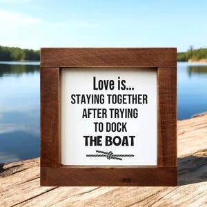 Op de afbeelding: Een vierkant houten bord met een donkerbruine lijst en een wit middenpaneel. De tekst luidt: Love is... STAYING TOGETHER AFTER TRYING TO DOCK THE BOAT. Onder de tekst staat een afbeelding van een touw in een knoop, alles in zwarte letters.