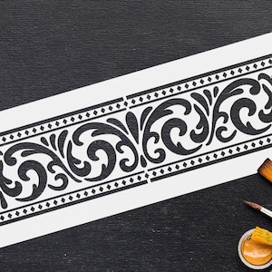 Könnte beinhalten: Eine schwarz-weiße Schablone mit einem dekorativen floralen Bordürendesign. Die Schablone befindet sich auf einem weißen Blatt Papier, mit Pinseln und Farbtöpfen im Hintergrund. Das Design umfasst Rautenformen und wirbelnde Blumenmuster.