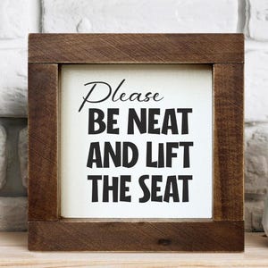 May include: A wooden framed sign with the text "Please BE NEAT AND LIFT THE SEAT" in black lettering. The sign is on a wooden shelf with a white vase to the right. The background is a white brick wall.
