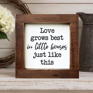 May include: A wooden framed sign with the text "Love grows best in little houses just like this" in black script. The sign is on a white background. A decorative wreath, a jar of flowers, and a metal pitcher are in the background.