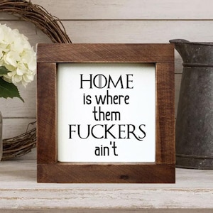 May include: A framed wooden sign with the text "HOME is where them FUCKERS ain't" in black lettering. The sign is set against a white background and framed in dark brown wood. Decorative home decor.
