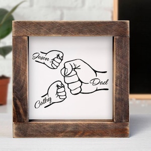 May include: A rustic wooden frame with a white background and a black and white illustration of three fists bumping together. The fists are labeled "Jason", "Cathy", and "Dad".