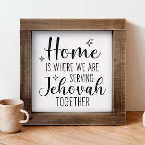 Może przedstawiać: Kwadratowy drewniany znak w ramie z napisem "Home is where we are serving Jehovah together" czarnym skryptem na białym tle. Widoczny jest ceramiczny kubek i inne elementy dekoracyjne.